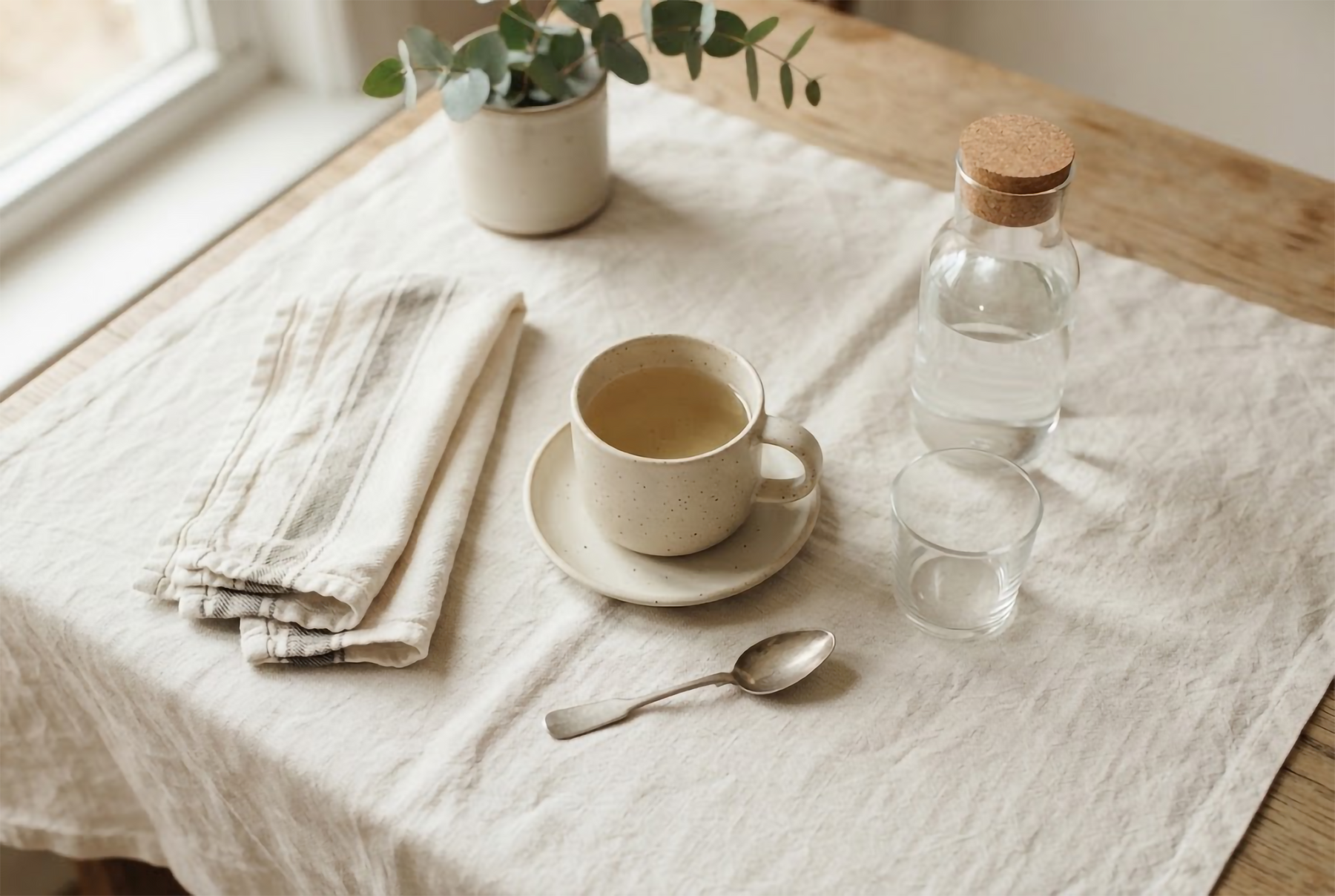 Morning routine flat-lay on ivory linen with ceramic cup and water carafe