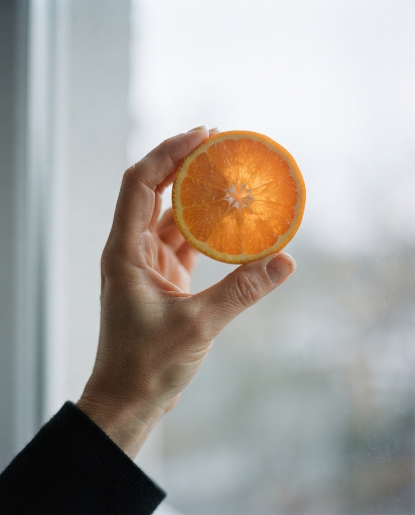 A hand holding a halved orange up to a bright window so the segments glow translucent.